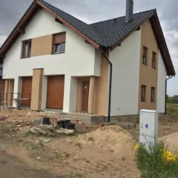 Dwukondygnacyjny budynek w trakcie prac elewacyjnych, z widoczną częścią otynkowaną na biało i beżowo, czarną dachówką, oraz niezagospodarowanym terenem wokół z gruzem i stertą piasku.