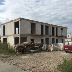 Surowy stan dwukondygnacyjnego budynku z betonu komórkowego w trakcie budowy, z widocznymi paletami, workami z materiałami budowlanymi i czerwonym vanem w tle.