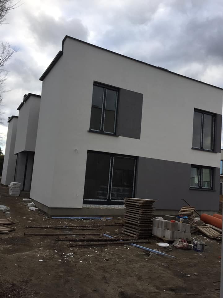 Nowoczesny dom z elewacją w odcieniach szarości, widoczne okna z ciemnymi ramami, teren wokół w trakcie prac wykończeniowych, stos palet i materiałów budowlanych.