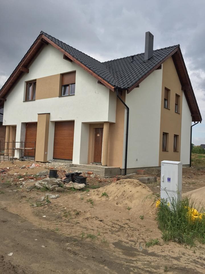 Dwukondygnacyjny budynek w trakcie prac elewacyjnych, z widoczną częścią otynkowaną na biało i beżowo, czarną dachówką, oraz niezagospodarowanym terenem wokół z gruzem i stertą piasku.