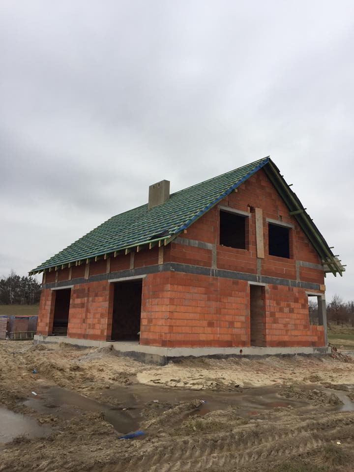 Surowy budynek z czerwonej cegły z zielonym dachem w trakcie budowy, widoczne niezabezpieczone otwory okienne i drzwiowe, otoczony błotnistym terenem.