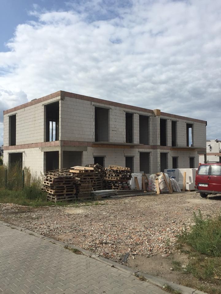 Surowy stan dwukondygnacyjnego budynku z betonu komórkowego w trakcie budowy, z widocznymi paletami, workami z materiałami budowlanymi i czerwonym vanem w tle.