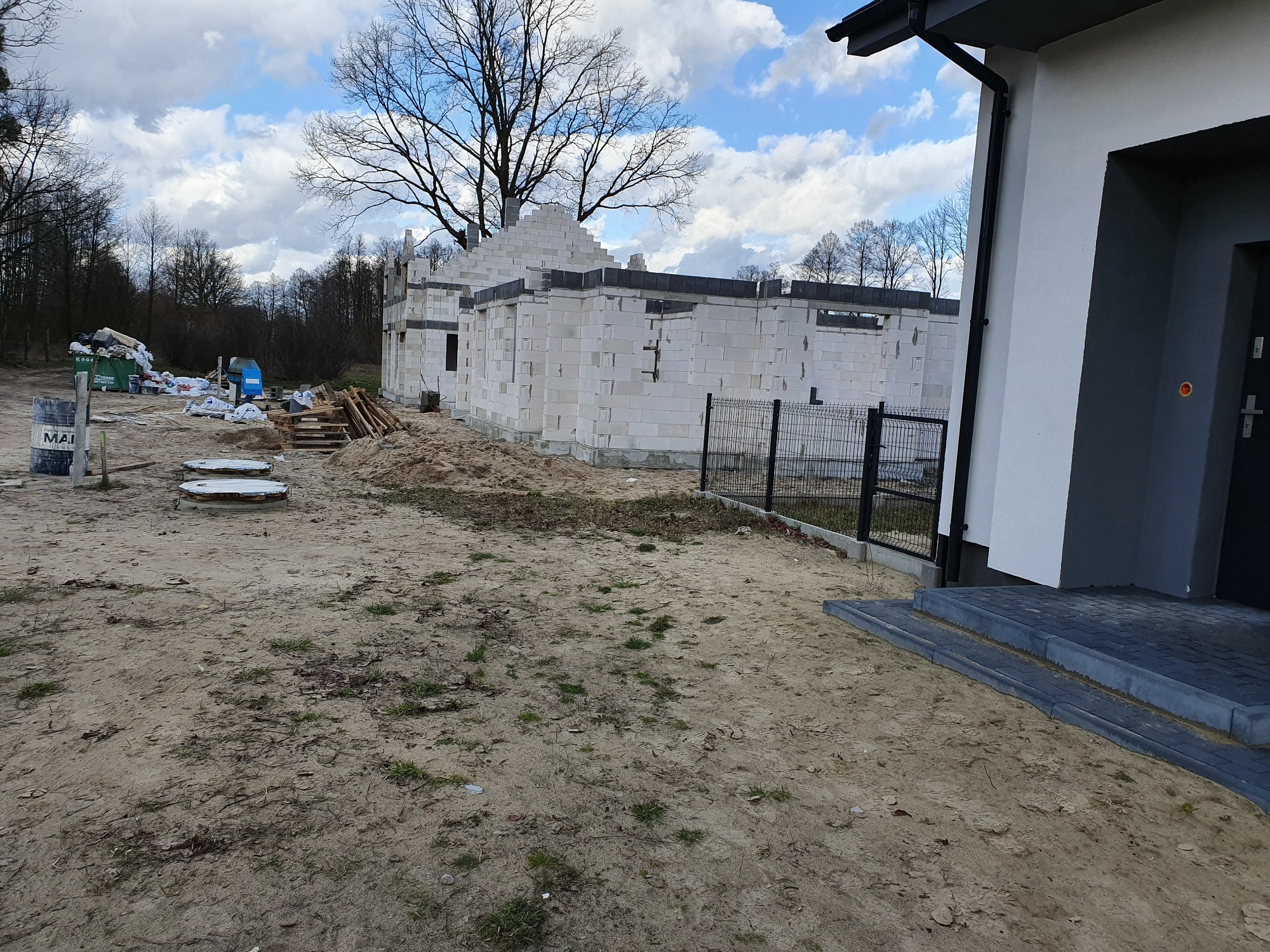 Budowa domu w stanie surowym otwartym z białego bloczka, widoczne fundamenty, ściany i elementy dachu, na pierwszym planie teren budowy z piaskiem, w tle drzewa i niebo z chmurami.