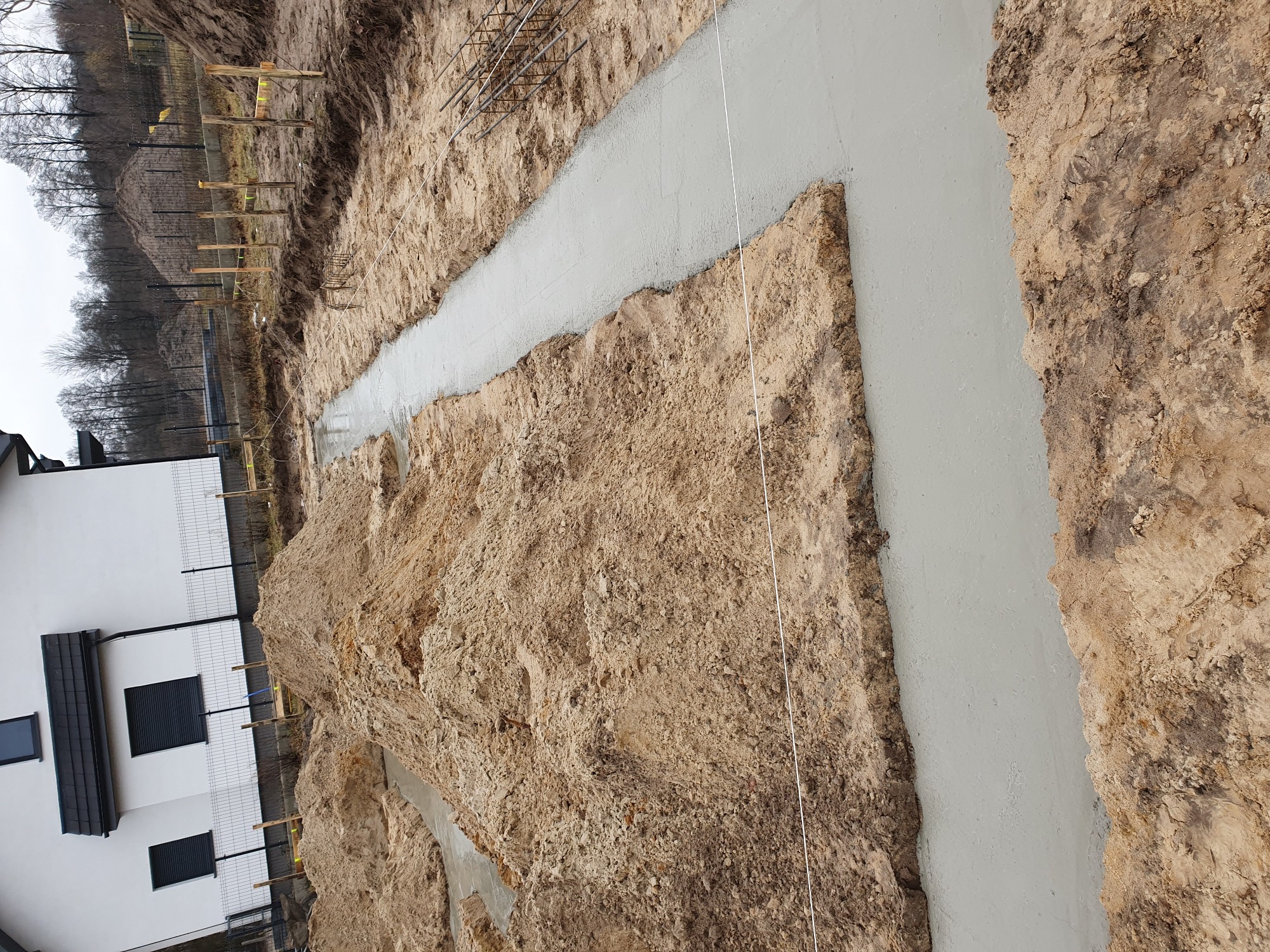 Wykop z zalanym betonem pod fundament domu, widoczne zbrojenie i dom w tle.