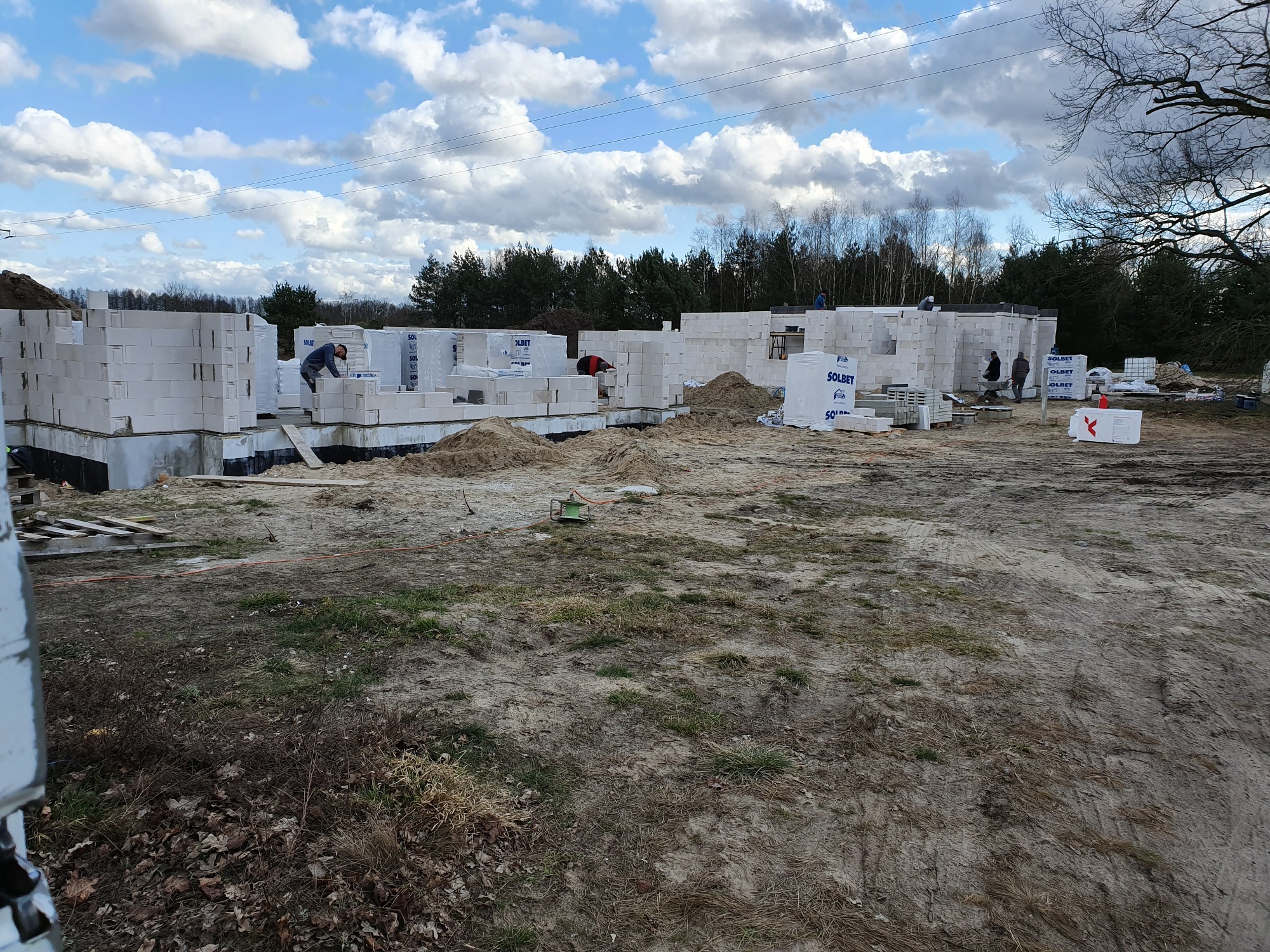 Budowa domu z bloczków betonu komórkowego na fundamencie, widoczne zapasy materiału Solbet, pracownicy murują ściany, w tle drzewa i pochmurne niebo.