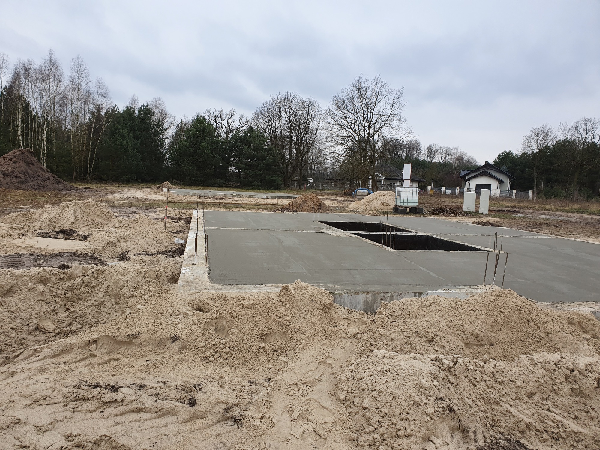 Wylany betonowy fundament z wystającymi prętami zbrojeniowymi, otoczony piaskiem, z widocznym wykopem pod instalacje, w tle drzewa i budynek gospodarczy.
