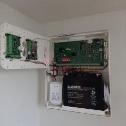 Otwarta obudowa centrali alarmowej z widocznymi płytkami drukowanymi, akumulatorem ALARMTEC i transformatorem, zamontowana na białej ścianie.
