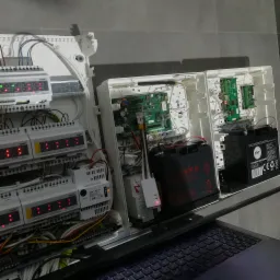 Rozdzielnica elektryczna z modułami sterującymi, płytkami drukowanymi i akumulatorami awaryjnymi w białych obudowach, widoczne diody LED sygnalizujące status, częściowo zasłonięta klawiatura laptopa.