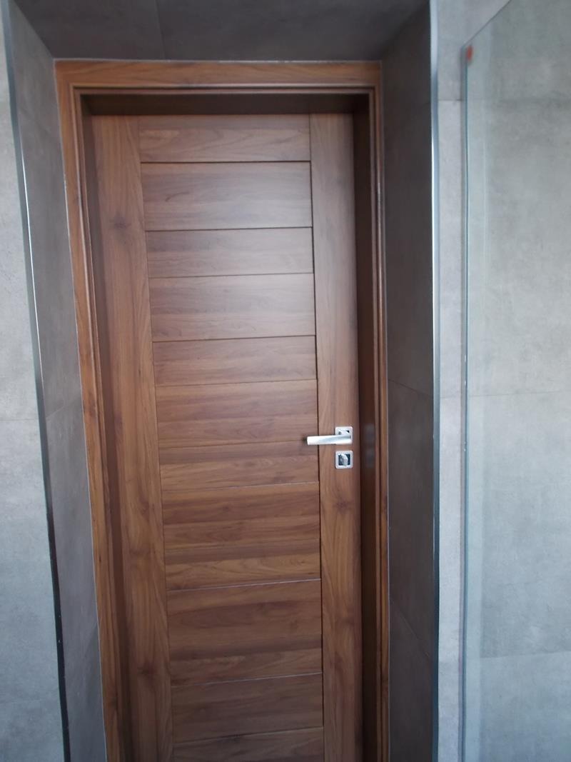 Zamknięte, drewniane skrzydło z poziomymi panelami w ościeżnicy, widoczne w łazience z szarą płytką i fragmentem kabiny prysznicowej.