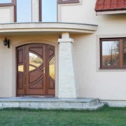 Wejście do domu z dwuskrzydłowymi, brązowymi drzwiami z dekoracyjnym przeszkleniem, wsparte na kolumnach, widok z zewnątrz w słoneczny dzień.
