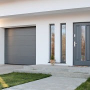 Nowoczesny dom jednorodzinny z szarą bramą garażową segmentową i szarymi drzwiami wejściowymi z przeszkleniami, widocznymi z zewnątrz na tle białej elewacji i trawnika.