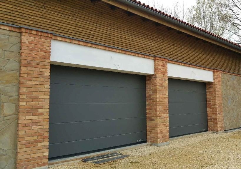 Dwa szare segmentowe zamknięcia garażowe z ceglanymi filarami i drewnianą elewacją, widok z zewnątrz.