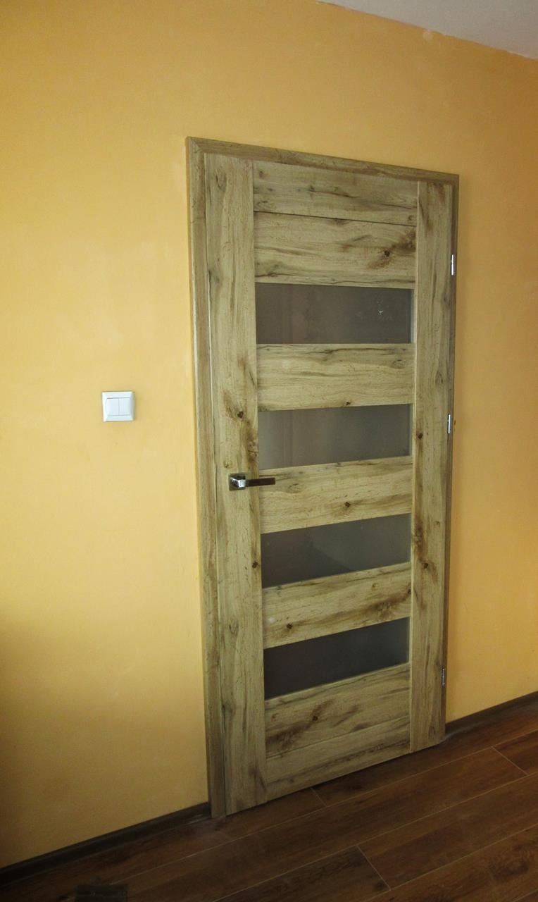 Drzwi wewnętrzne Invado z drewnopodobną okleiną i czterema poziomymi przeszkleniami, zamontowane w ścianie o żółtym odcieniu.