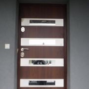 Ciemnobrązowe skrzydło z poziomymi przeszkleniami i srebrnymi elementami osprzętu na tle szarej ściany.