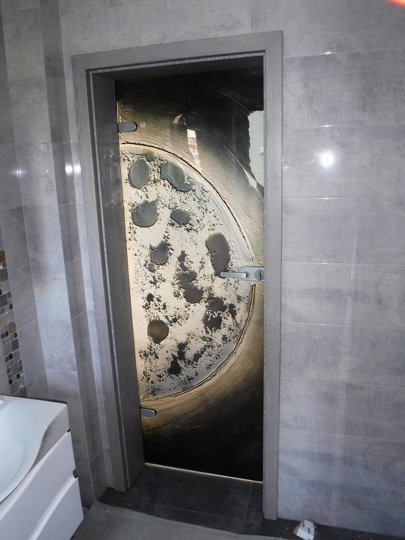 Szklana tafla z abstrakcyjnym wzorem przypominającym kratery, zamontowana jako element oddzielający pomieszczenia, widoczna klamka i szara ościeżnica.