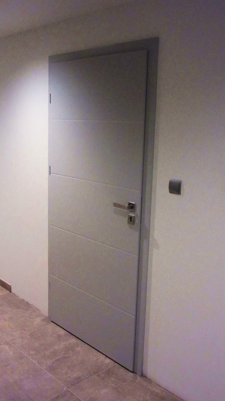 Szare, nowoczesne skrzydło z poziomymi frezami i srebrną klamką, zamontowane w jasnym wnętrzu z szarą podłogą z płytek.