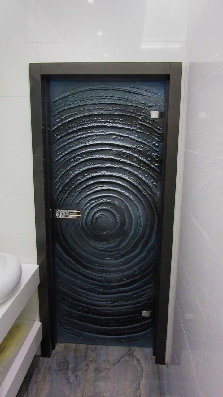 Ciemna, nowoczesna konstrukcja z wzorem koncentrycznych okręgów, osadzona w ciemnej ramie, z widocznym zamkiem i zawiasami, wkomponowana w łazienkę z białymi płytkami i marmurową podłogą.