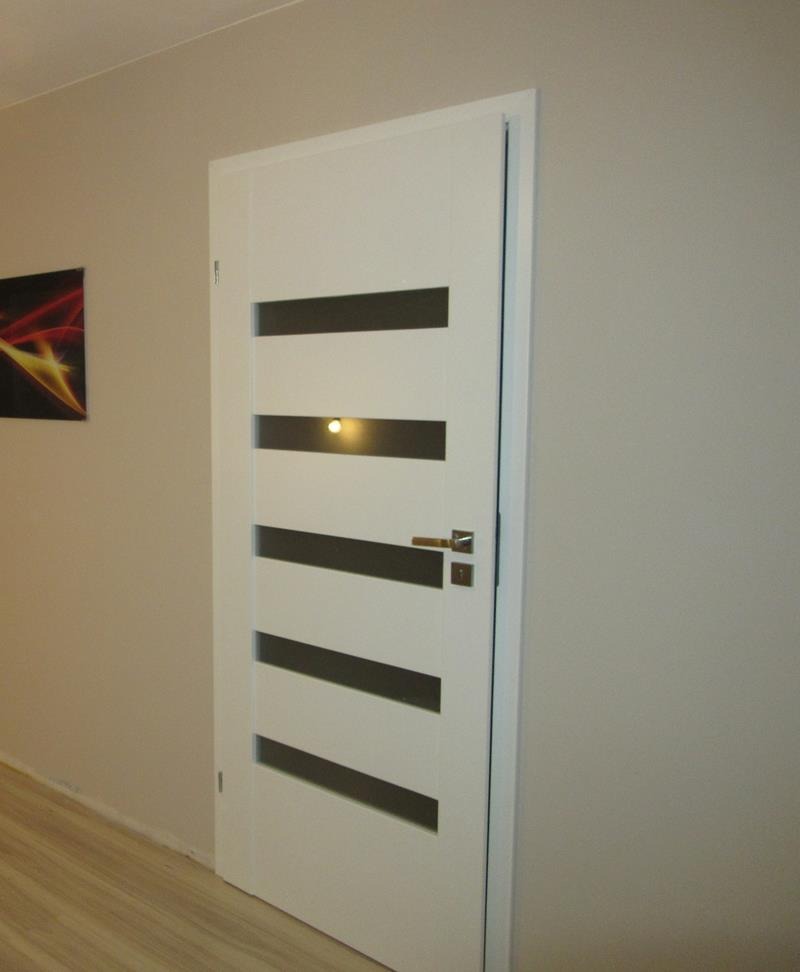 Białe skrzydło z czterema poziomymi przeszkleniami, zamontowane w jasnym wnętrzu, widoczna klamka i zamek.