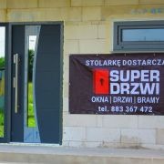Szare wejście do domu z zamontowanymi nowoczesnymi drzwiami z przeszkleniem, obok widoczny baner reklamowy firmy 'Super Drzwi' oferującej stolarkę okienną, drzwi i bramy.