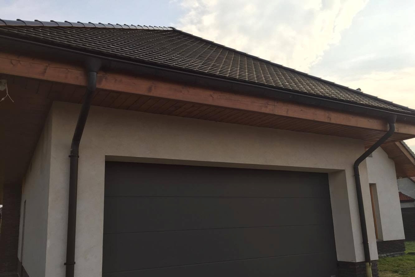 Szara segmentowa brama garażowa z poziomymi przetłoczeniami, zamontowana w elewacji budynku z brązowym dachem i rynnami