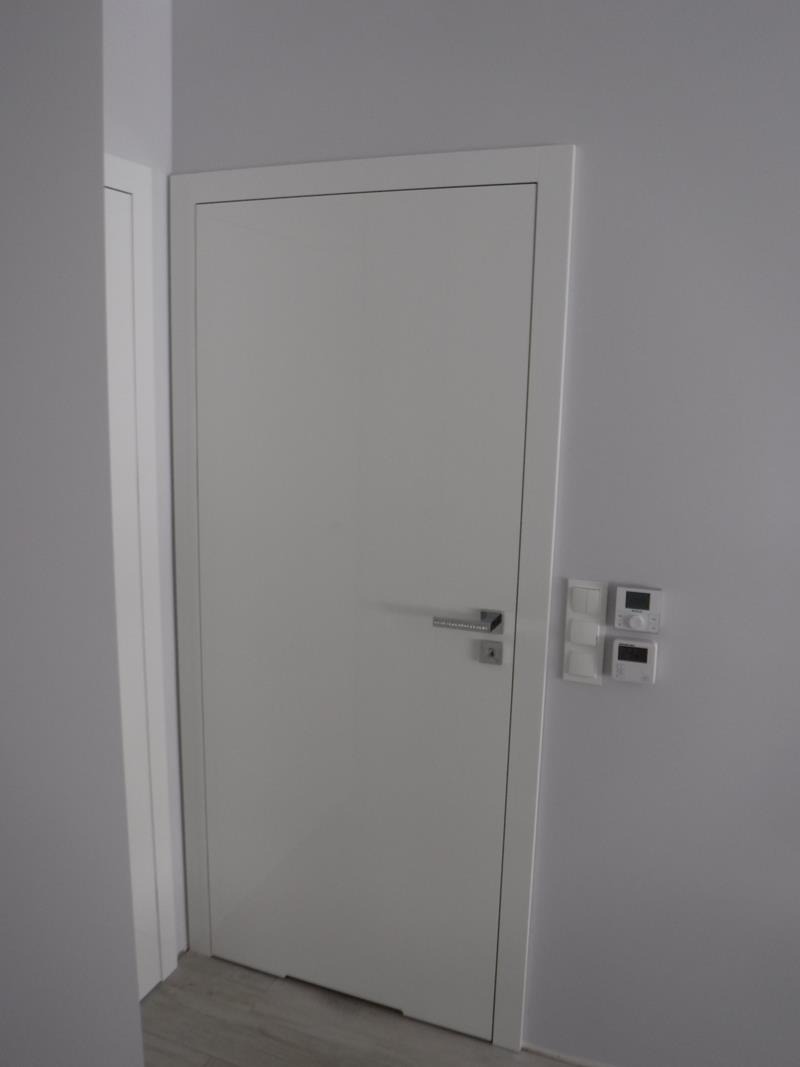 Białe skrzydło z ościeżnicą w minimalistycznym wnętrzu, widoczny chromowany klamka i elektroniczne panele sterowania na ścianie.