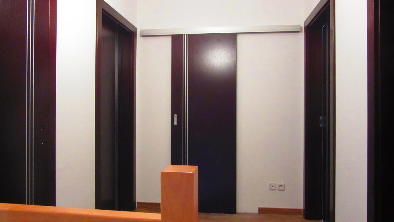 Ciemnobrązowe skrzydło przesuwne z trzema srebrnymi paskami, widoczne na tle białej ściany, obok standardowych otwieranych skrzydeł w ciemnej okleinie, z elementami konstrukcyjnymi w kolorze...