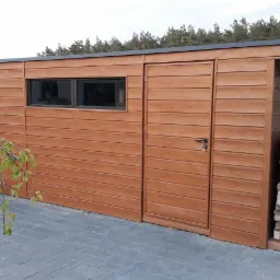 Nowoczesna, drewniana konstrukcja z oknem i drzwiami, przylegająca do niej wiata na drewno opałowe, na tle kostki brukowej i domu.