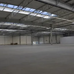 Przestronna hala przemysłowa z szarą posadzką, widoczne stalowe słupy, oświetlenie sufitowe i brama wjazdowa w tle. Posadzka gładka, bez widocznych uszkodzeń.