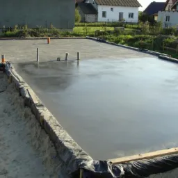 Świeżo wylana posadzka na fundamencie, z widocznymi pionowymi rurami i deskowaniem. Tło stanowią budynki mieszkalne i trawa.