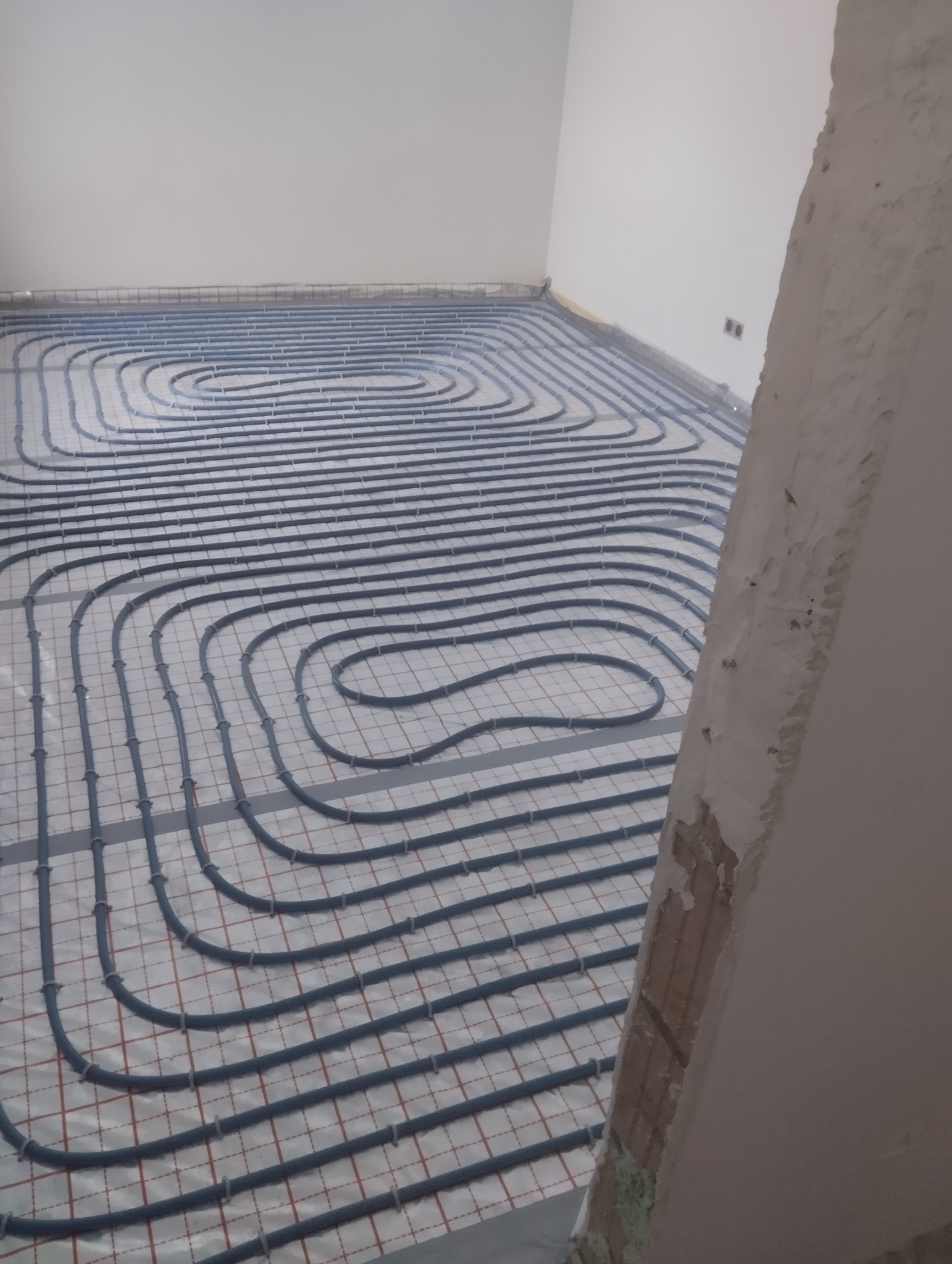 Rozłożona instalacja ogrzewania podłogowego z szarych rurek na folii z siatką, widok z góry, surowy stan wykończenia pomieszczenia z fragmentem obdrapanej ściany w prawym dolnym rogu.