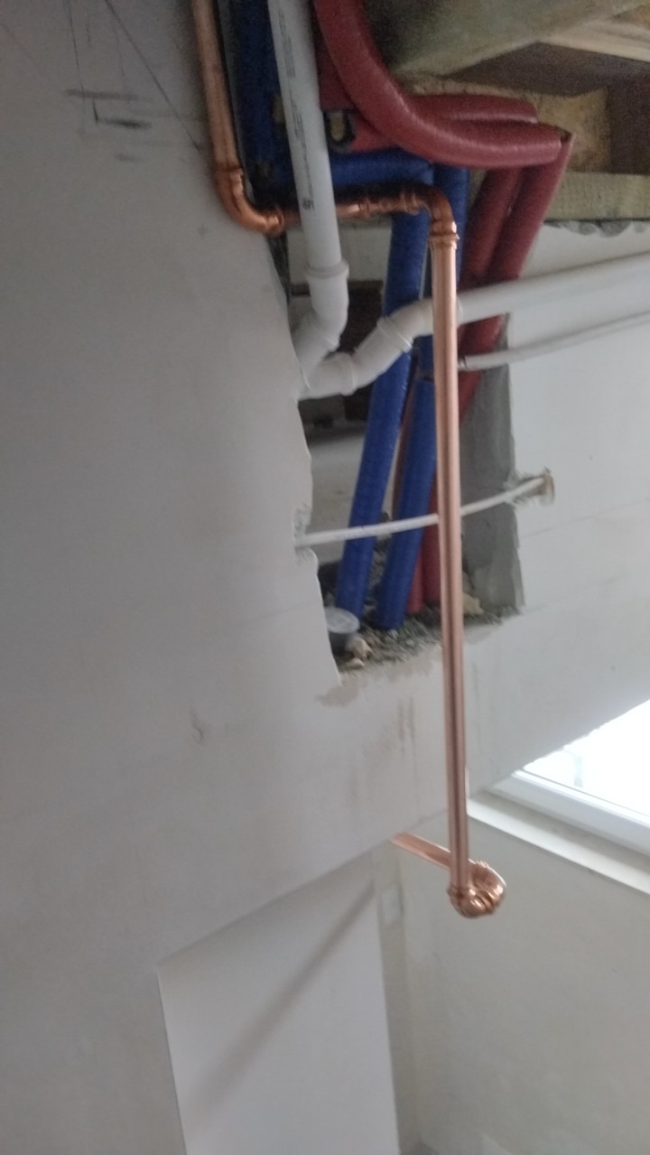 Instalacja hydrauliczna w trakcie montażu: rury miedziane, białe i niebieskie rury plastikowe widoczne w otworze w ścianie, surowy tynk.