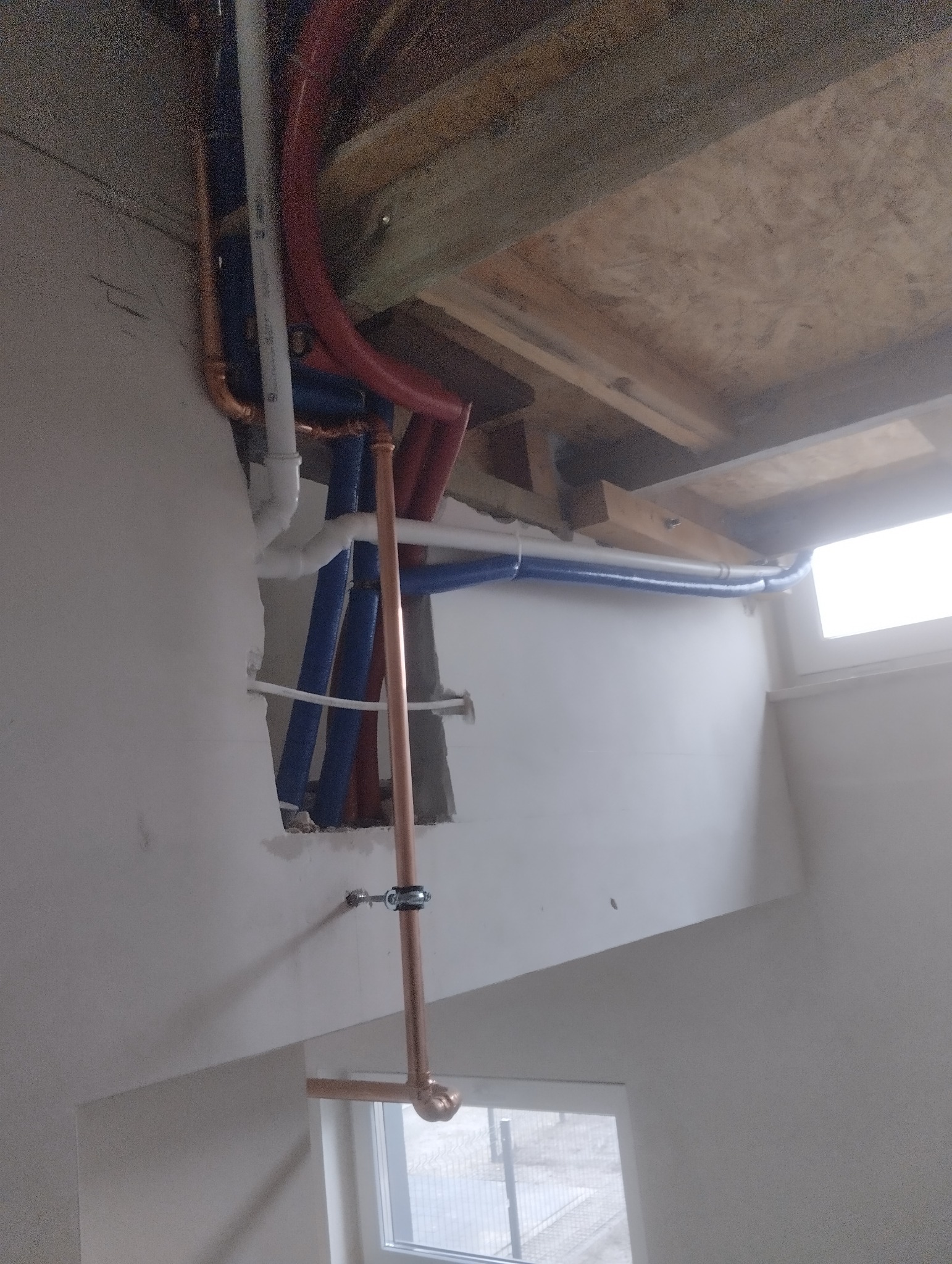 Wnętrze w trakcie instalacji wodno-kanalizacyjnej: widoczne pionowe i poziome rury miedziane, plastikowe rury w kolorze niebieskim i czerwonym, mocowane do ściany i pod stropem. Częściowo...