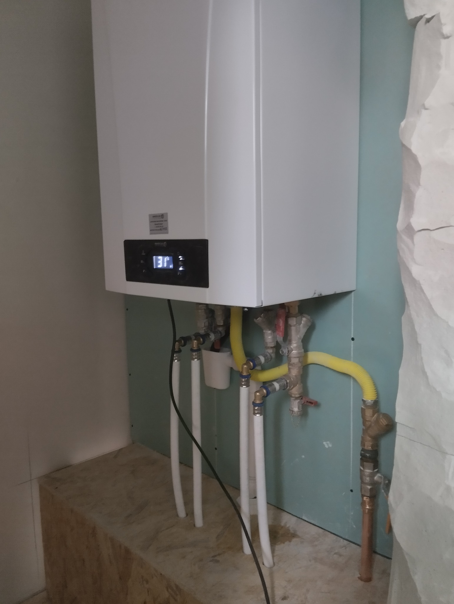 Biały piec gazowy wiszący na ścianie z podłączonymi rurami wodnymi i gazowymi, widoczny wyświetlacz z temperaturą, tło stanowią surowe ściany i podłoga w trakcie wykończenia.