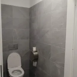 Nowoczesna toaleta z szarymi, wielkoformatowymi płytkami imitującymi kamień, wisząca muszla klozetowa i metalowy podajnik na papier toaletowy.