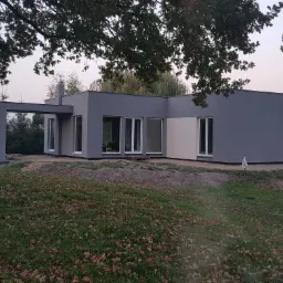 Nowoczesny, parterowy dom z płaskim dachem, pomalowany na szaro, z dużymi oknami, widoczny w otoczeniu drzew i trawnika z opadłymi liśćmi.