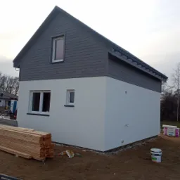 Dwukondygnacyjny budynek w trakcie wykańczania elewacji, z drewnianą boazerią na górnej kondygnacji pomalowaną na szaro i jasną, gładką elewacją na dole, z widocznymi oknami, stertą drewna...