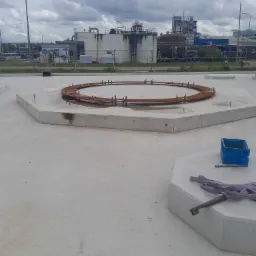 Widok z góry na przygotowany betonowy fundament pod przemysłowy zbiornik, z widocznym zardzewiałym pierścieniem zbrojeniowym i elementami montażowymi w otoczeniu przemysłowym.