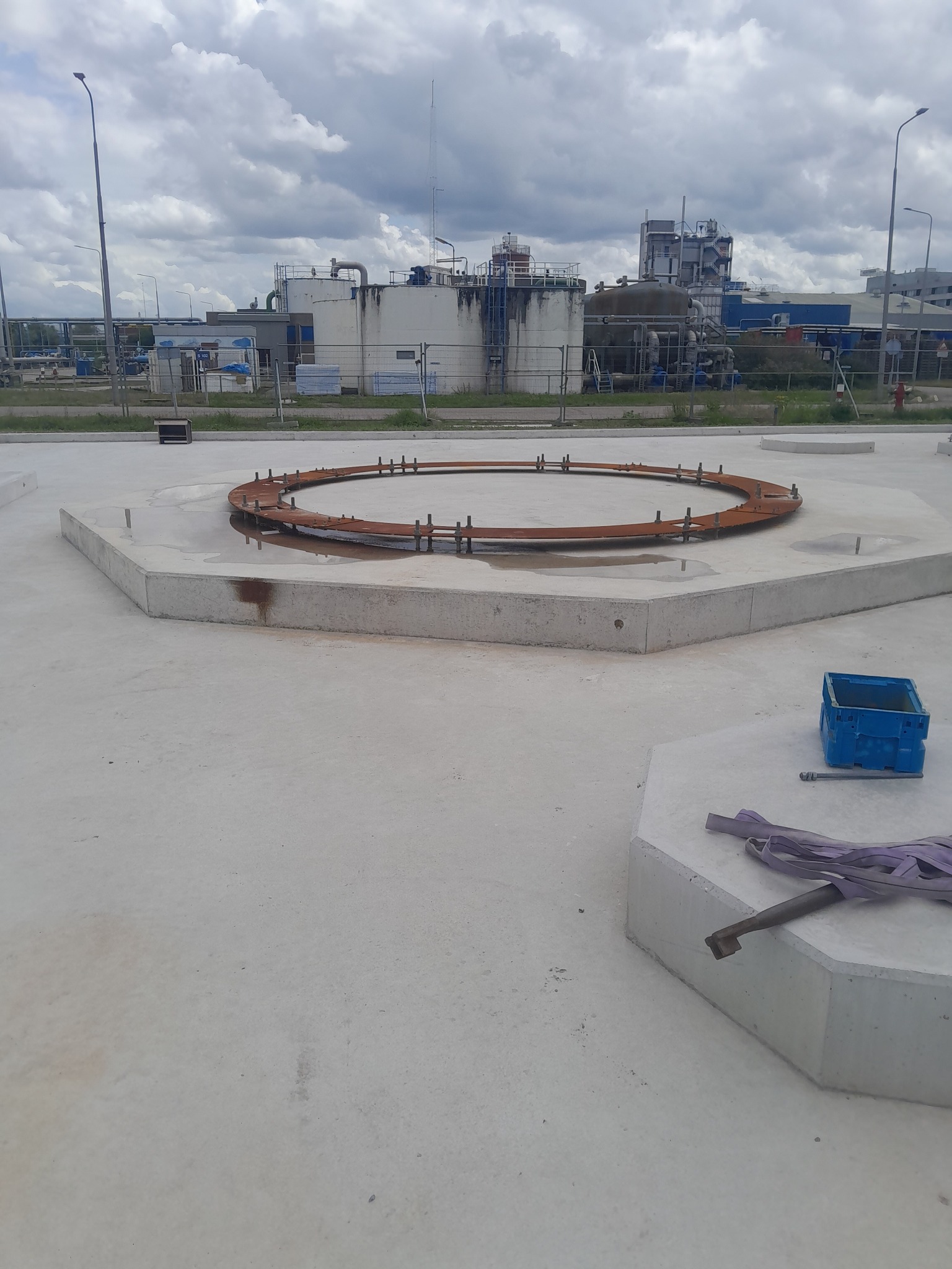 Widok z góry na przygotowany betonowy fundament pod przemysłowy zbiornik, z widocznym zardzewiałym pierścieniem zbrojeniowym i elementami montażowymi w otoczeniu przemysłowym.