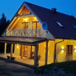 Dwukondygnacyjny dom z drewna z balkonem i zadaszonym tarasem oświetlony ciepłym światłem w scenerii zmierzchu. Na dachu widoczne okna dachowe i antena satelitarna.