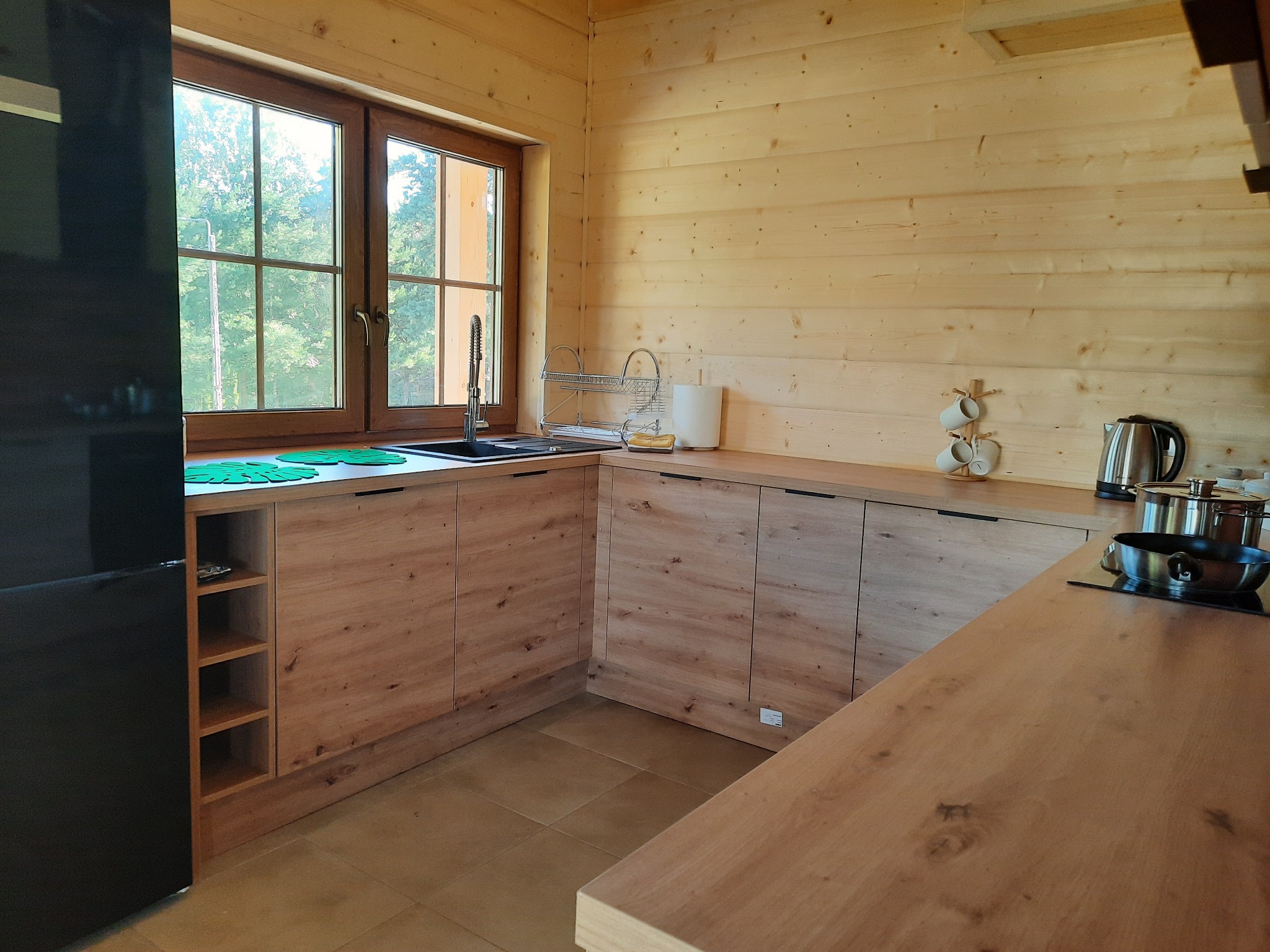 Jasna, drewniana kuchnia w domu szkieletowym z widokiem na zieleń za oknem, wyposażona w nowoczesne szafki z drewnianymi frontami i czarnymi uchwytami, oraz czarną lodówkę.