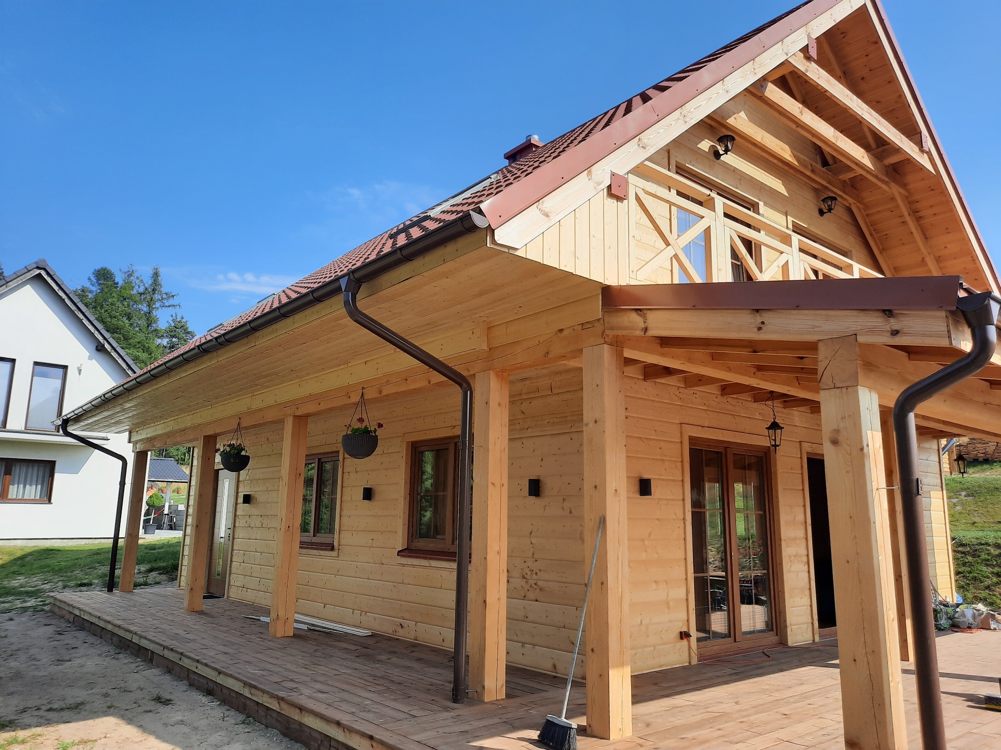 Nowoczesny dom z jasnego drewna z tarasem i balkonem, widoczne detale wykończenia dachu i orynnowania w kolorze brązowym, na pierwszym planie zamiataczka.
