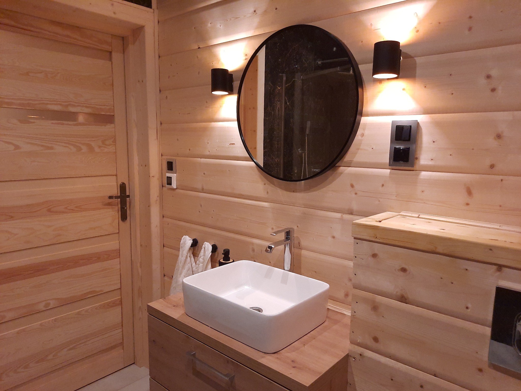 Wnętrze łazienki w domu z drewnianych bali, z okrągłym lustrem, białą umywalką nablatową i drewnianymi meblami, oświetlone ciepłym światłem kinkietów.