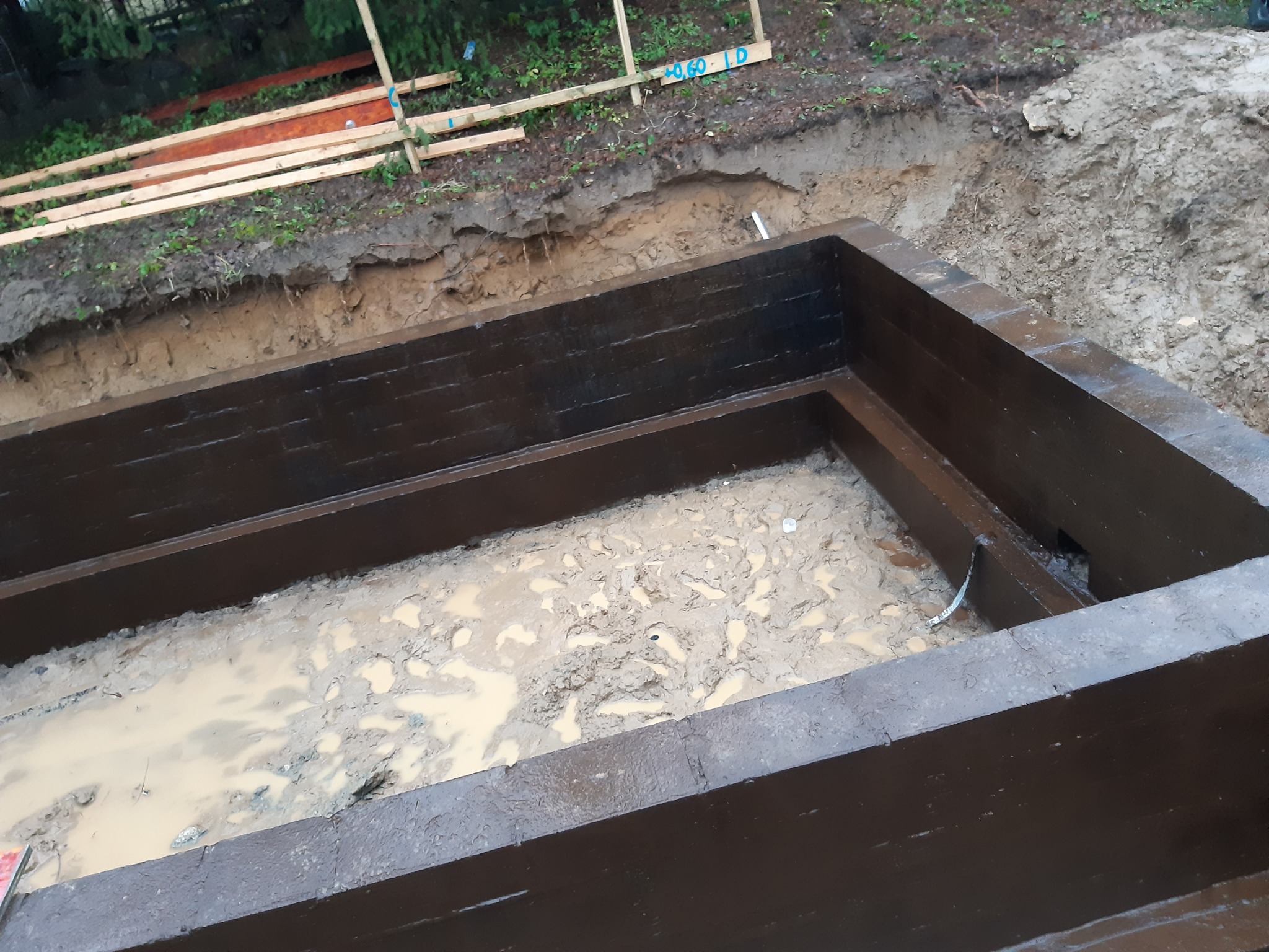 Wykop pod fundament z zalanym wodą błotem, zabezpieczony deskami z pomalowaną na ciemno impregnacją, widoczne drewniane stemple i oznaczenia geodezyjne na krawędzi wykopu.