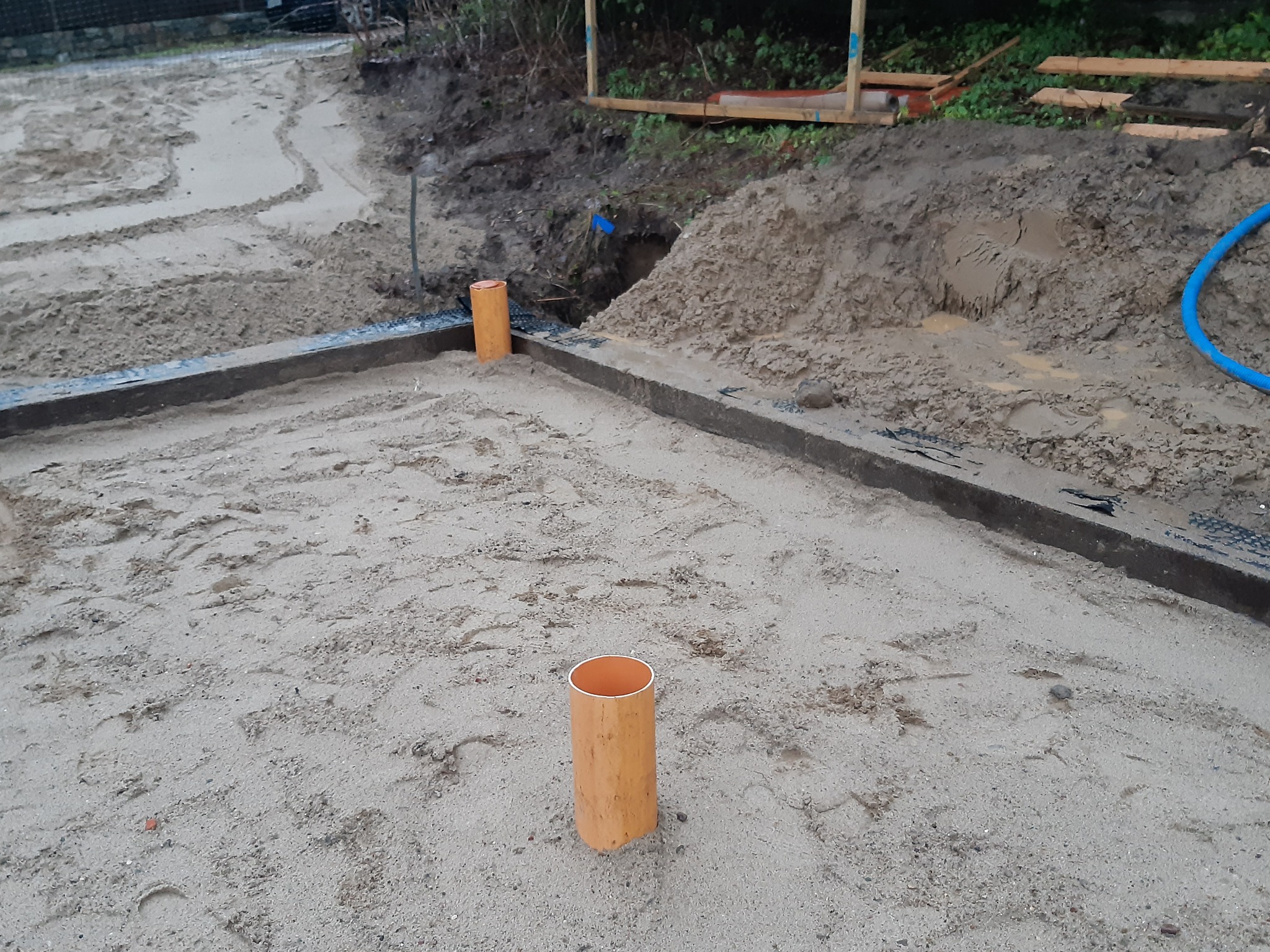 Przygotowanie podłoża pod wylewkę: piasek wyrównany, obrzeża z szalunków, widoczne pomarańczowe rury instalacyjne, ziemia i niebieski wąż w tle.
