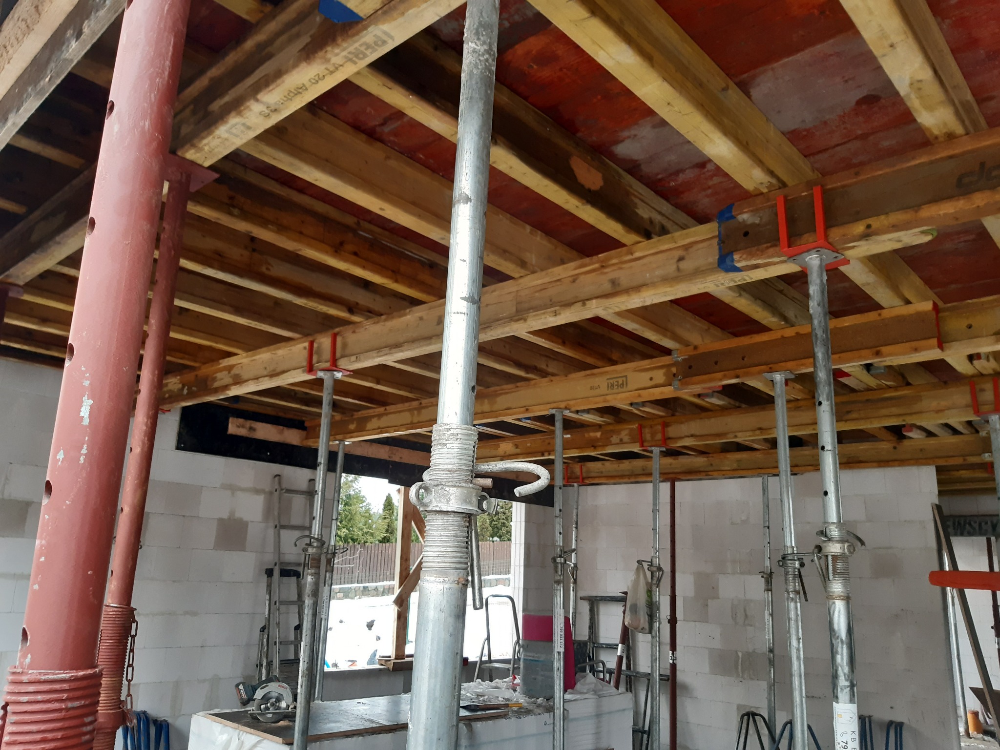 Szare, teleskopowe stemple budowlane podtrzymujące drewniany szalunek stropu na budowie domu z białych bloczków.