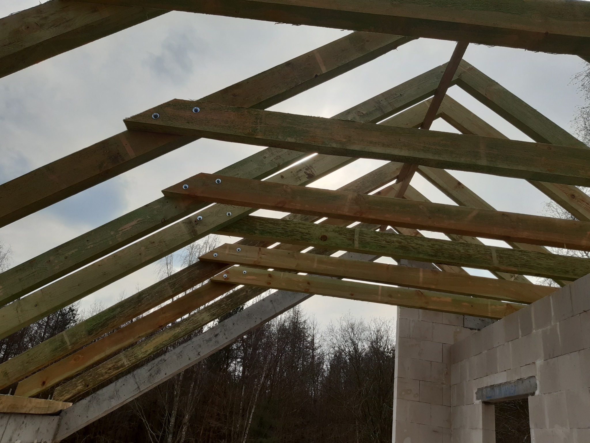 Konstrukcja dachu z drewnianych belek, zabezpieczonych śrubami, na tle nieba i częściowo wybudowanych ścian z betonu komórkowego.