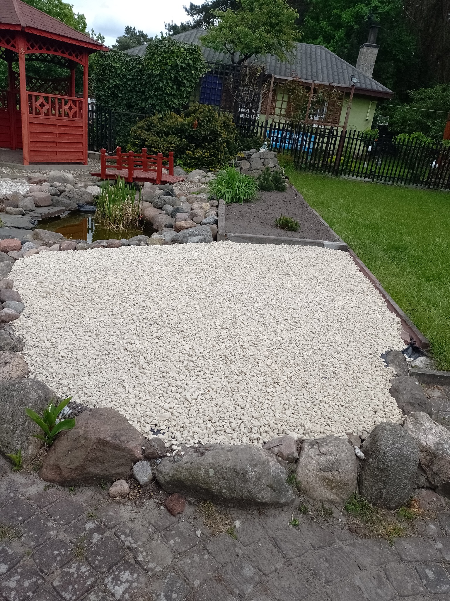 Ogród z oczkiem wodnym i wysypaną białą korą kamienną, otoczony kamieniami. W tle altana i dom z zielenią. Całość tworzy harmonijny krajobraz.