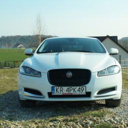 Sebastian Kołodziejczyk Tiases Taxi - Biały samochód Jaguar XF z tablicą rejestracyjną KR 4PK49, zaparkowany na żwirowej nawierzchni przed wiejskim krajobrazem z budynkami i polami.