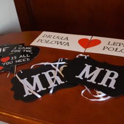 Sebastian Kołodziejczyk Tiases Taxi - Rekwizyty do fotobudki na brązowym stole: czarne tabliczki z napisami 'MR' na białych wstążkach, 'I DANC. I'M QUISIN', 'HE', 'I CAME FOR THE IS ALL YOU NEED' z czerwonym sercem, oraz kartki...