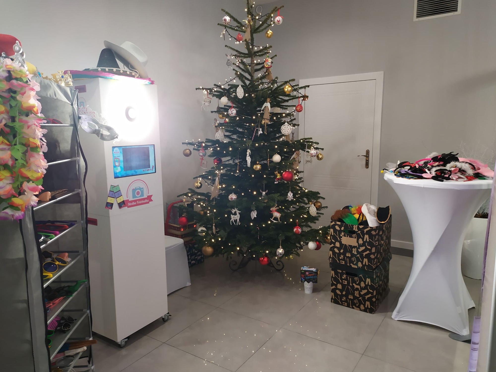 Fotobudka z ekranem dotykowym i rekwizytami w świątecznej scenerii z choinką, pudełkami prezentowymi i stolikiem z przebraniami.